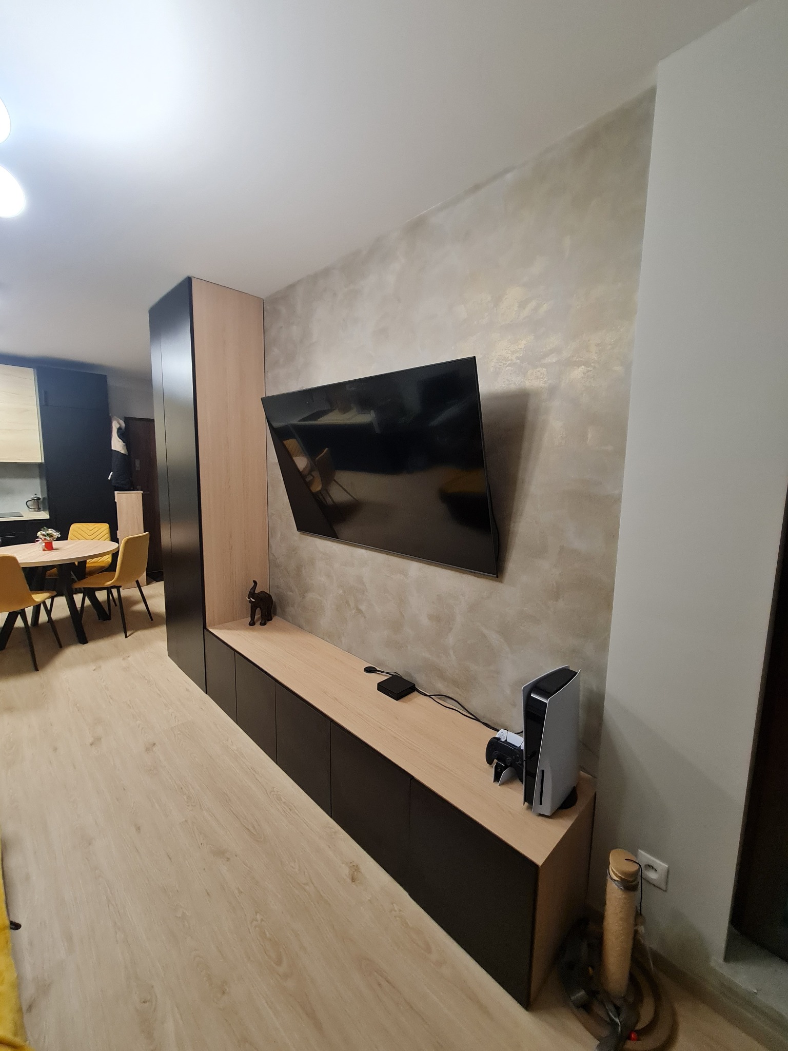 Nowoczesna szafka RTV z czarnymi frontami i drewnianym blatem, na ścianie telewizor, obok konsola PlayStation 5, w tle widoczna kuchnia z żółtymi krzesłami.
