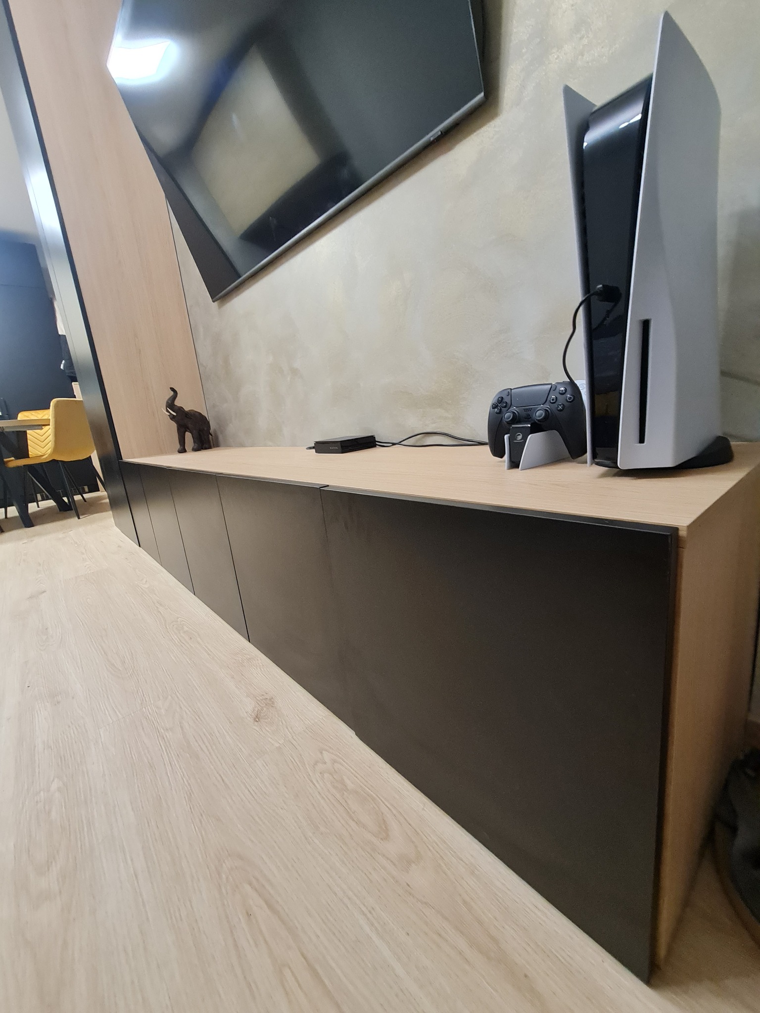 Nowoczesna szafka RTV z czarnymi frontami i drewnianym blatem, na której stoi konsola PlayStation 5 z kontrolerem, oraz dekoracyjna figurka słonia. W tle widoczny telewizor.