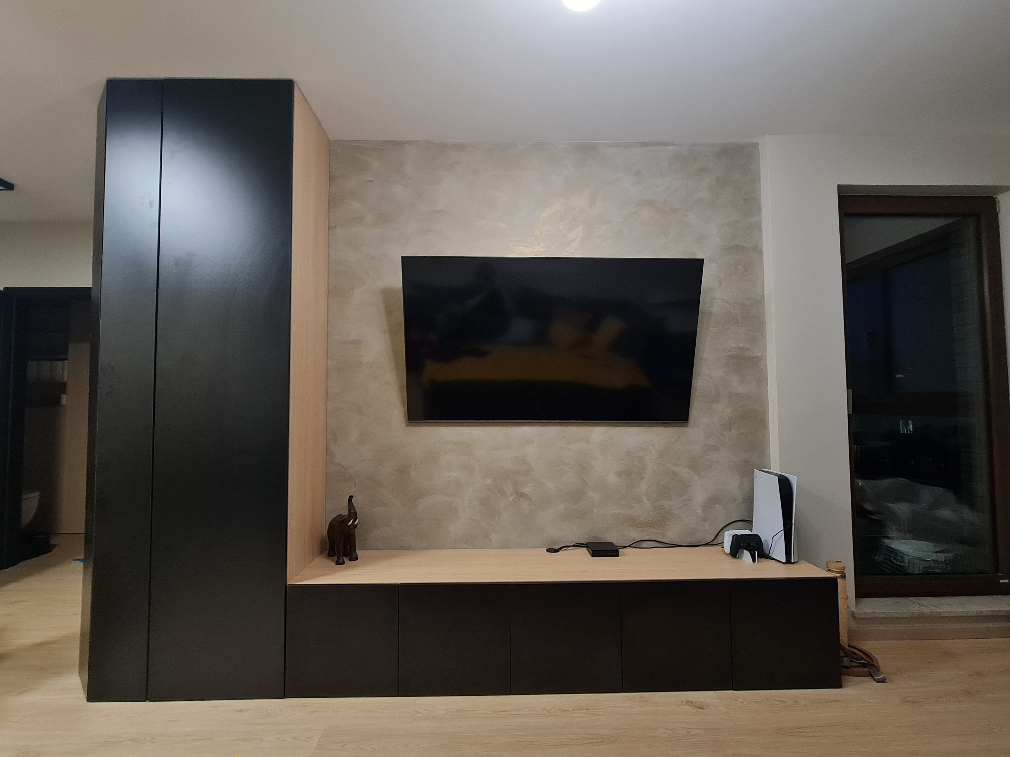 Czarno-beżowa zabudowa meblowa z telewizorem, konsolą PlayStation 5 i figurką słonia na blacie, na tle ściany z efektem dekoracyjnym.