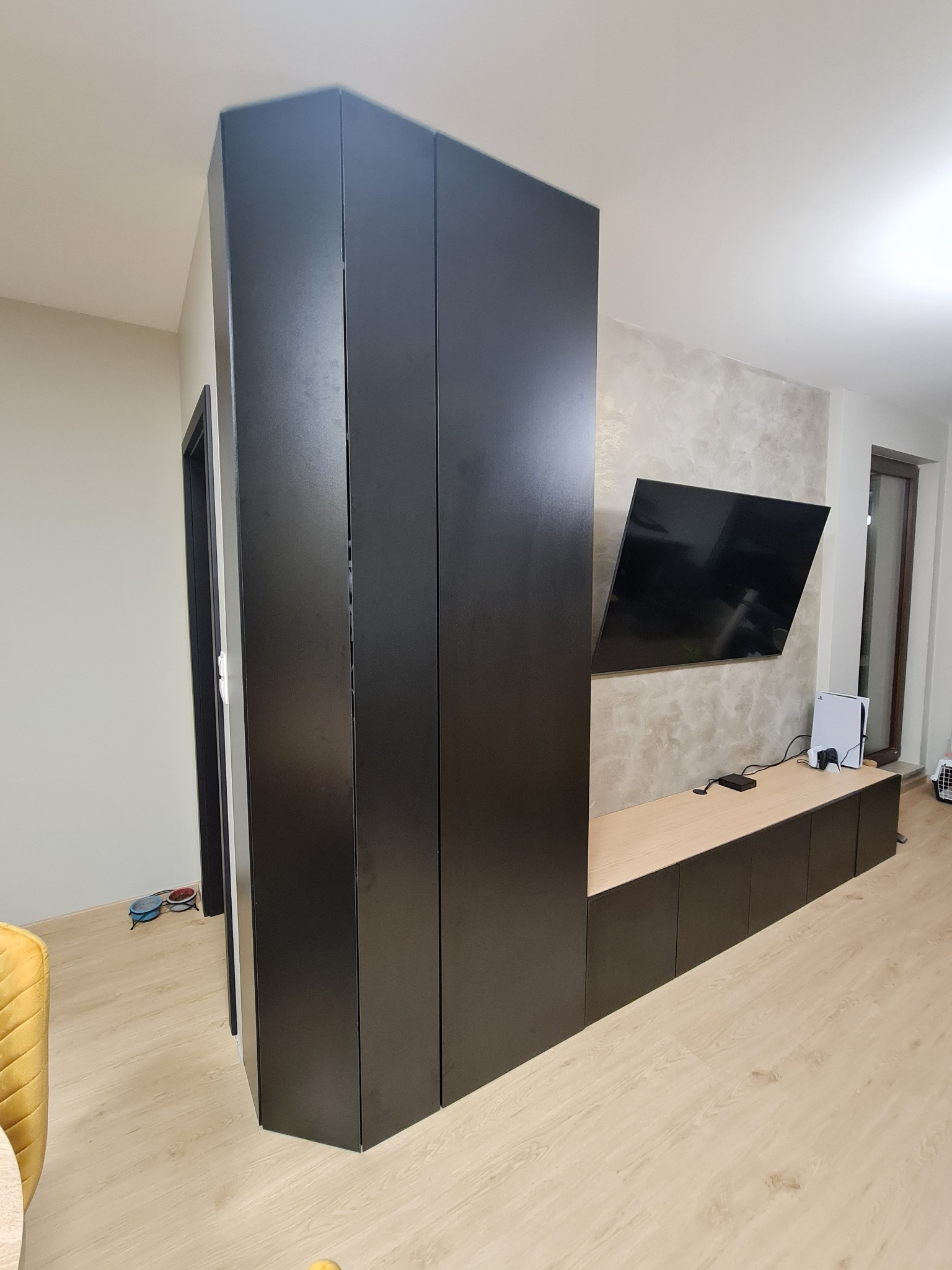 Czarna zabudowa meblowa dzieląca przestrzeń z szafami i szafkami RTV, telewizor na ścianie o fakturze betonu, jasna podłoga.