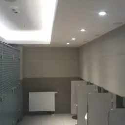 Publiczna toaleta z kabinami o wzorzystych ściankach, szare płytki ścienne, biały grzejnik i oświetlenie sufitowe.