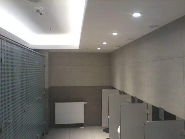 Publiczna toaleta z kabinami o wzorzystych ściankach, szare płytki ścienne, biały grzejnik i oświetlenie sufitowe.