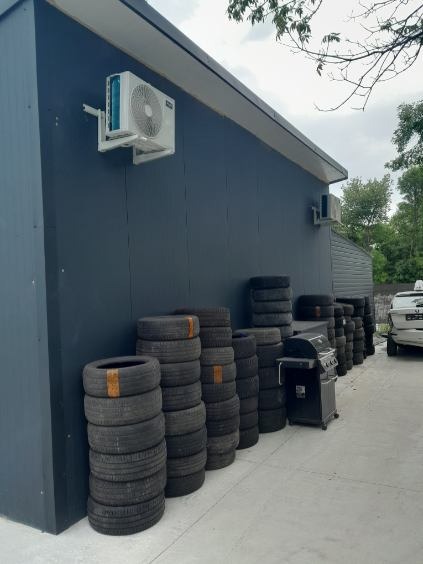 Zewnętrzna jednostka klimatyzacji zamontowana na ciemnej elewacji budynku, obok stosy opon i grill na zewnątrz.