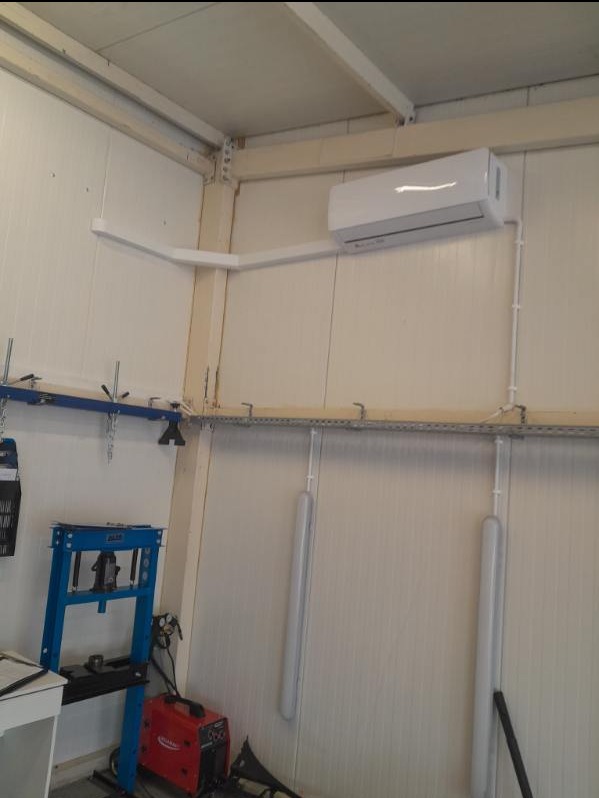 Biały klimatyzator ścienny zamontowany w pomieszczeniu warsztatowym, widoczne rury instalacyjne, prasa hydrauliczna i spawarka.