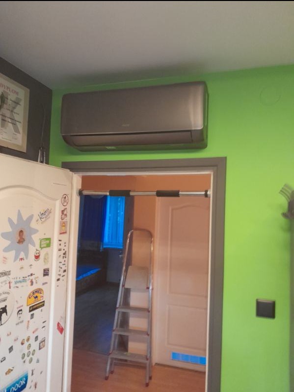 Szary klimatyzator zamontowany nad futryną drzwi, widoczna drabina oparta o ścianę, ściana pomalowana na jaskrawo zielony kolor, na ścianie wisi dyplom.
