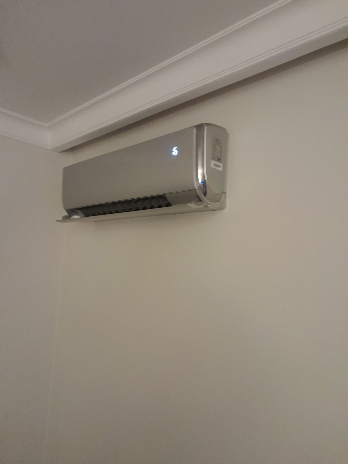 Srebrny klimatyzator ścienny z wyświetlaczem temperatury na białej ścianie z ozdobnym gzymsem sufitowym.