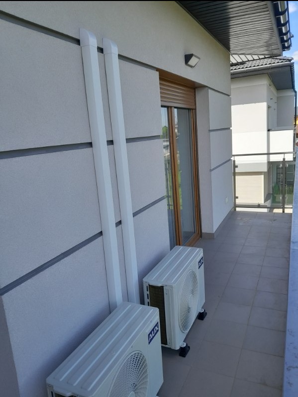 Dwa zainstalowane zewnętrzne moduły klimatyzacji marki AUX na balkonie budynku mieszkalnego, z widocznymi białymi rurami odprowadzającymi, zamontowane na szarych podstawkach antywibracyjnych.