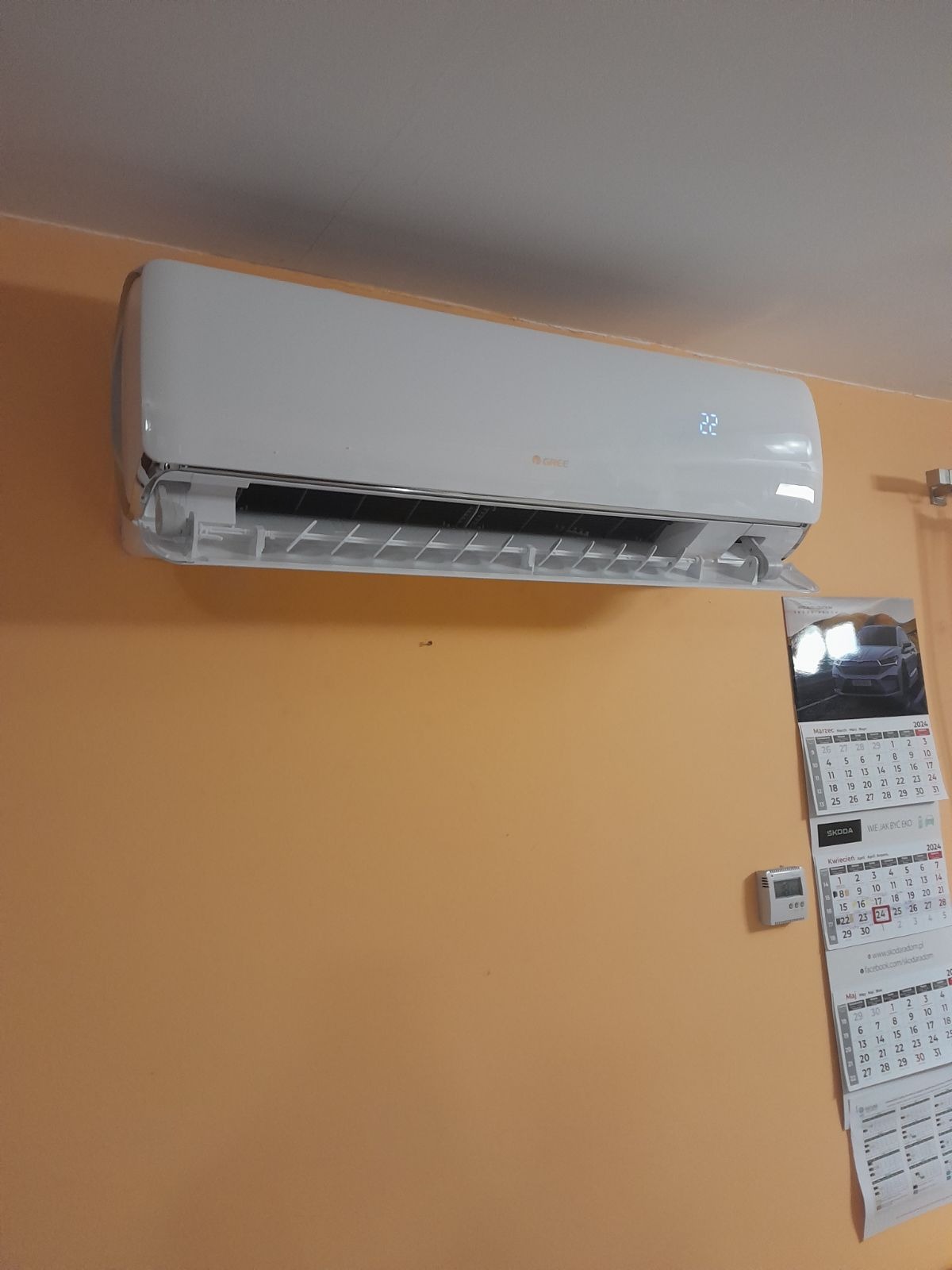 Biała klimatyzacja ścienna marki Gree, ustawiona na 22 stopnie, zamontowana na pomarańczowej ścianie w tle widoczny kalendarz.