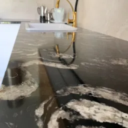 Czarny blat z białymi żyłami, z widocznym złotym kranem i naczyniami kuchennymi w tle.