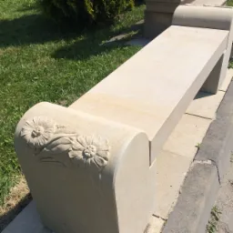 Beżowa ławka z kamienia z rzeźbionymi kwiatami na boku, ustawiona na chodniku obok trawnika.