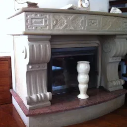 Obudowa kominka z piaskowca z dekoracyjnymi detalami, wykończona blatem z czerwonego granitu, z widoczną wazą ceramiczną i drewnianymi schodami w tle.