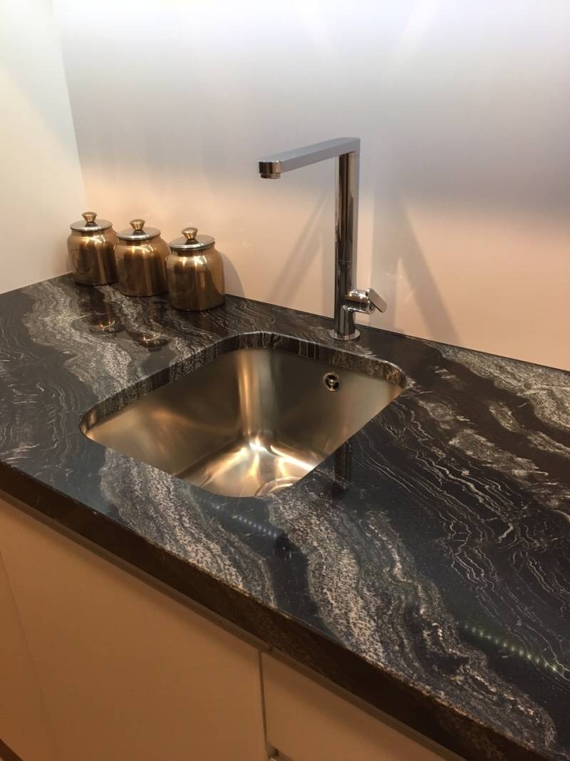 Polerowany blat z czarnego granitu z białymi żyłkami, z wpuszczonym zlewozmywakiem ze stali nierdzewnej i nowoczesną baterią, obok trzy złote pojemniki.