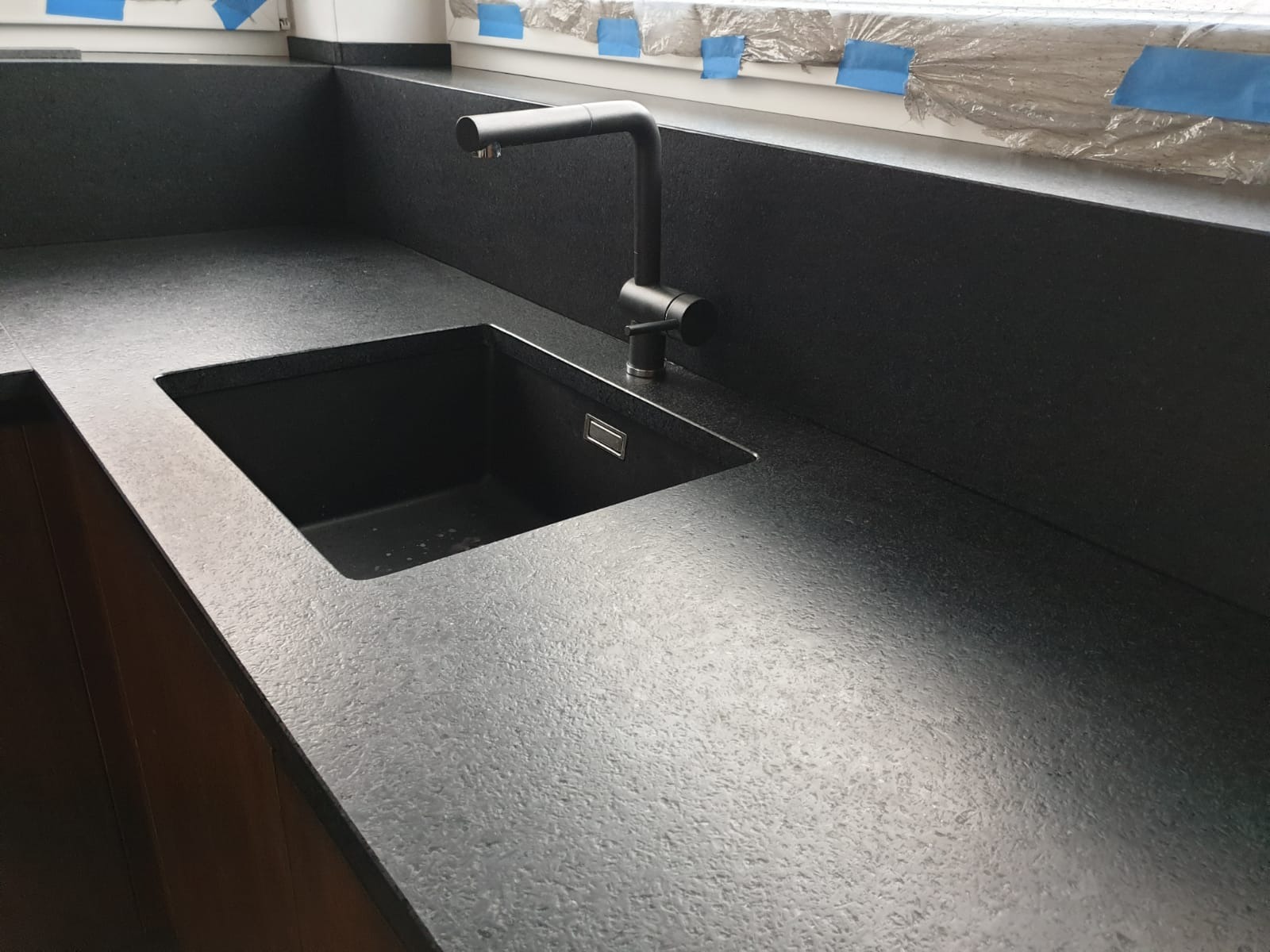 Czarny, matowy blat z umywalką i nowoczesną baterią w kuchni, okno zabezpieczone folią malarską w tle.