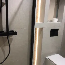 Nowoczesna łazienka z kabiną prysznicową typu walk-in, podświetlanymi niszami w ścianie i podwieszaną toaletą. Wykończenie w jasnych płytkach.