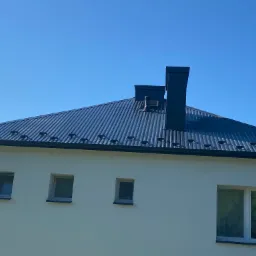 Nowoczesny dom z ciemnoszarą, metalową blachodachówką, widoczne okna i komin na tle błękitnego nieba.