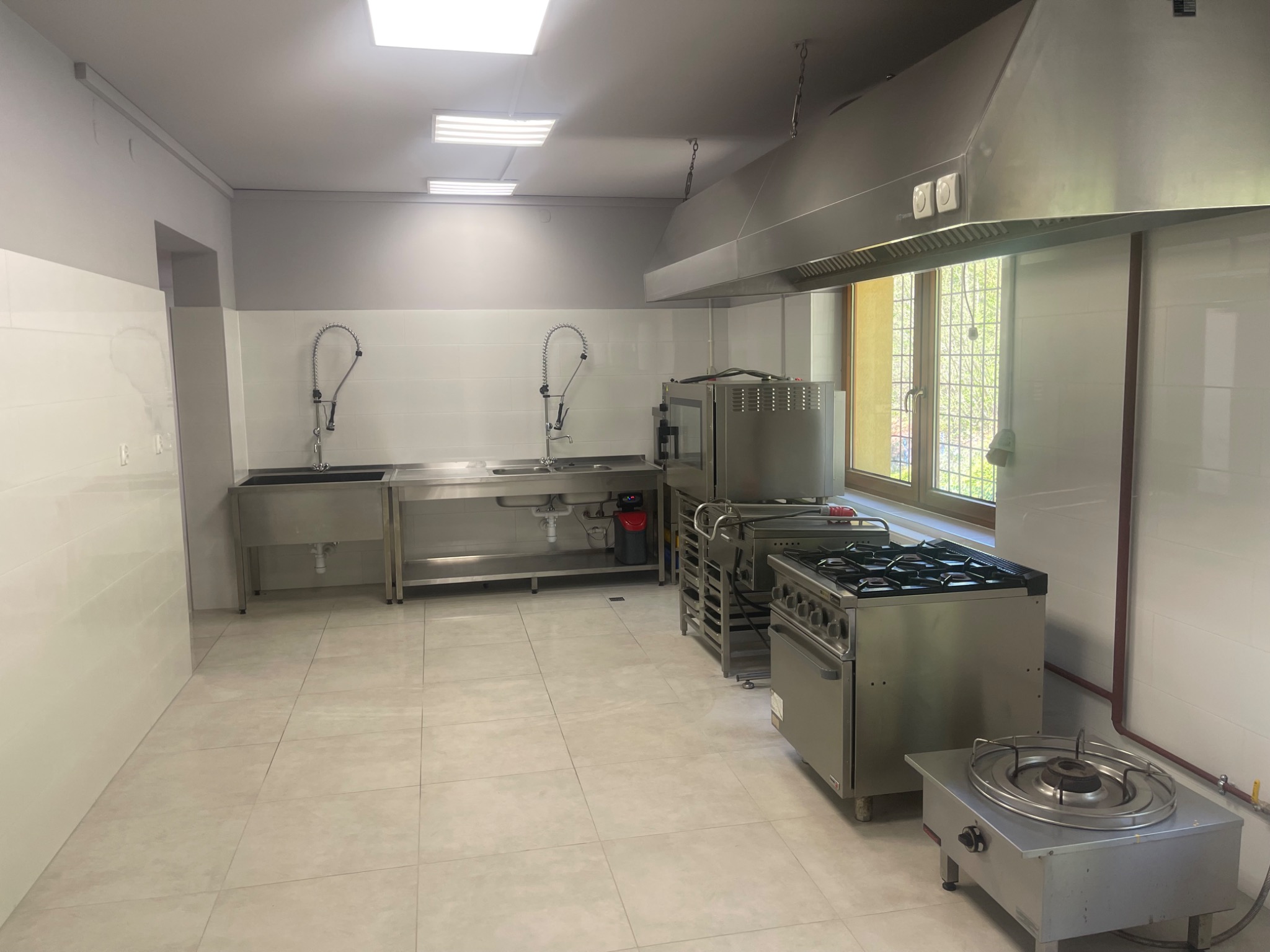 Wyposażona kuchnia przemysłowa ze stali nierdzewnej: zlewozmywaki z bateriami, okap, piekarnik i kuchenka gazowa. Białe płytki na ścianach i podłodze. Jasne, naturalne światło.