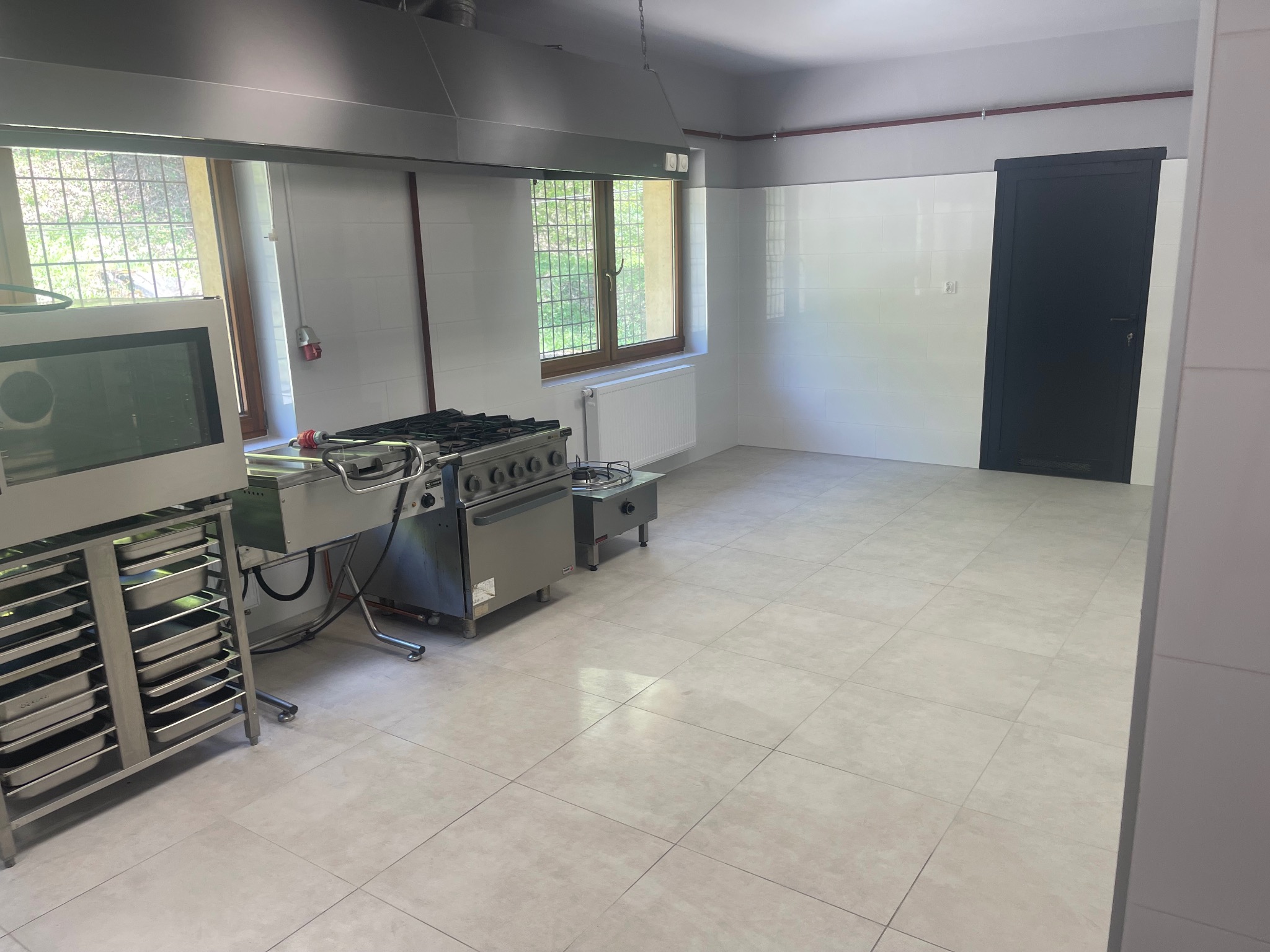 Wyposażona kuchnia przemysłowa: stalowy piec gazowy z okapem, regał na blachy i białe kafelki na ścianach. Widok na okno i drzwi.