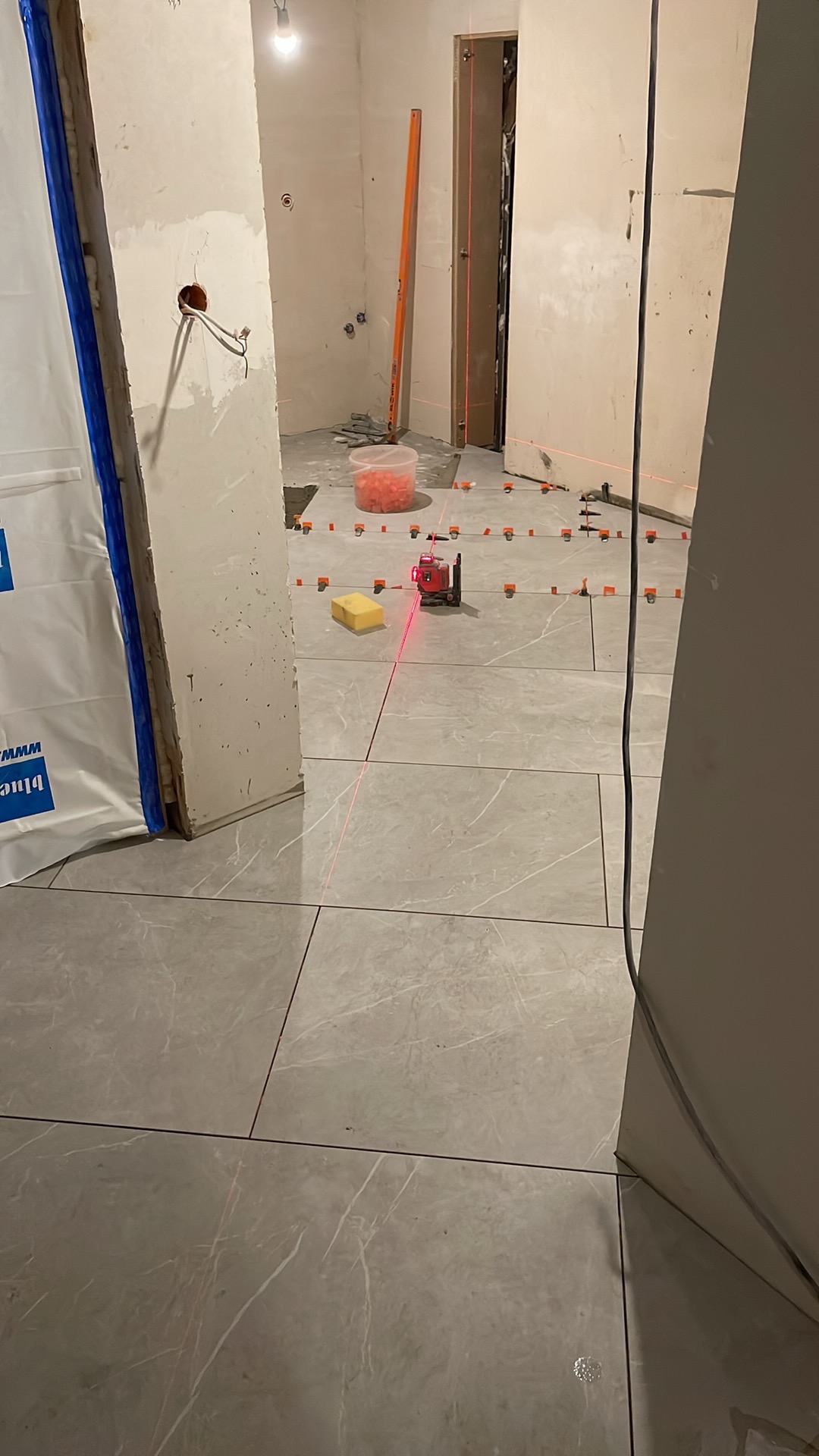 Układanie płytek podłogowych z użyciem poziomicy laserowej. Widoczne krzyżyki dystansowe i wiadro z zaprawą. Częściowo wykończona podłoga w odcieniach szarości.