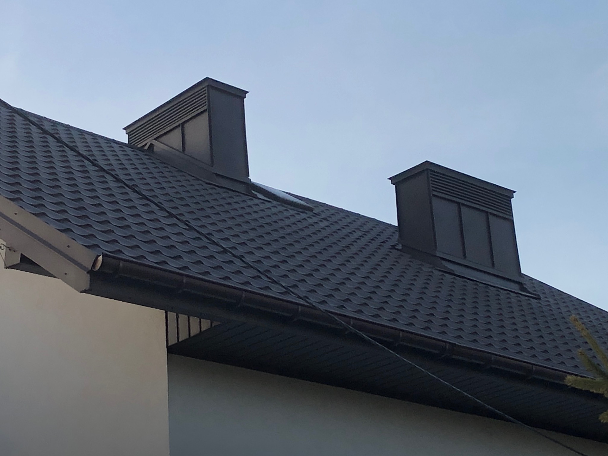 Grafitowy dach z dwoma nowoczesnymi kominami wentylacyjnymi na tle jasnego nieba. Widok z dołu, akcentujący teksturę dachówki i minimalistyczny design kominów.