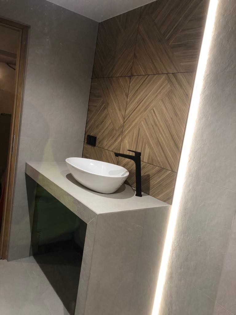 Biała umywalka nablatowa z czarną armaturą na szarym blacie, na tle drewnopodobnych płytek ułożonych we wzór. Oświetlenie LED wzdłuż ściany.