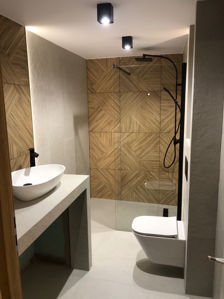 Nowoczesna łazienka z drewnianą mozaiką na ścianie prysznica, białą umywalką nablatową i podwieszaną toaletą. Minimalistyczne oświetlenie sufitowe.