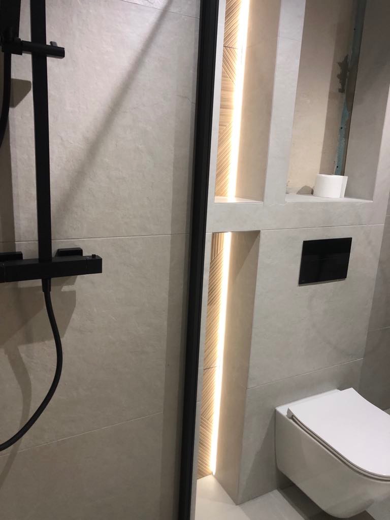 Nowoczesna łazienka z kabiną prysznicową typu walk-in, podświetlanymi niszami w ścianie i podwieszaną toaletą. Wykończenie w jasnych płytkach.