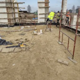 Budowa domu: robotnicy w odblaskowych kamizelkach montują ściany z bloczków na fundamencie, widoczne szalunki, zbrojenia, piła łańcuchowa na ziemi.