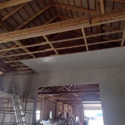 Wnętrze budynku w trakcie prac budowlanych: widoczne drewniane krokwie dachu, częściowo zamontowane białe panele sufitowe, rusztowania, worki z materiałem budowlanym i wózek widłowy w tle.