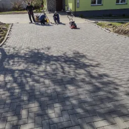 Układanie szarej kostki brukowej na podjeździe przed domem z zieloną elewacją, trzech pracowników w trakcie pracy, zagęszczarka obok, widoczne cienie drzew.