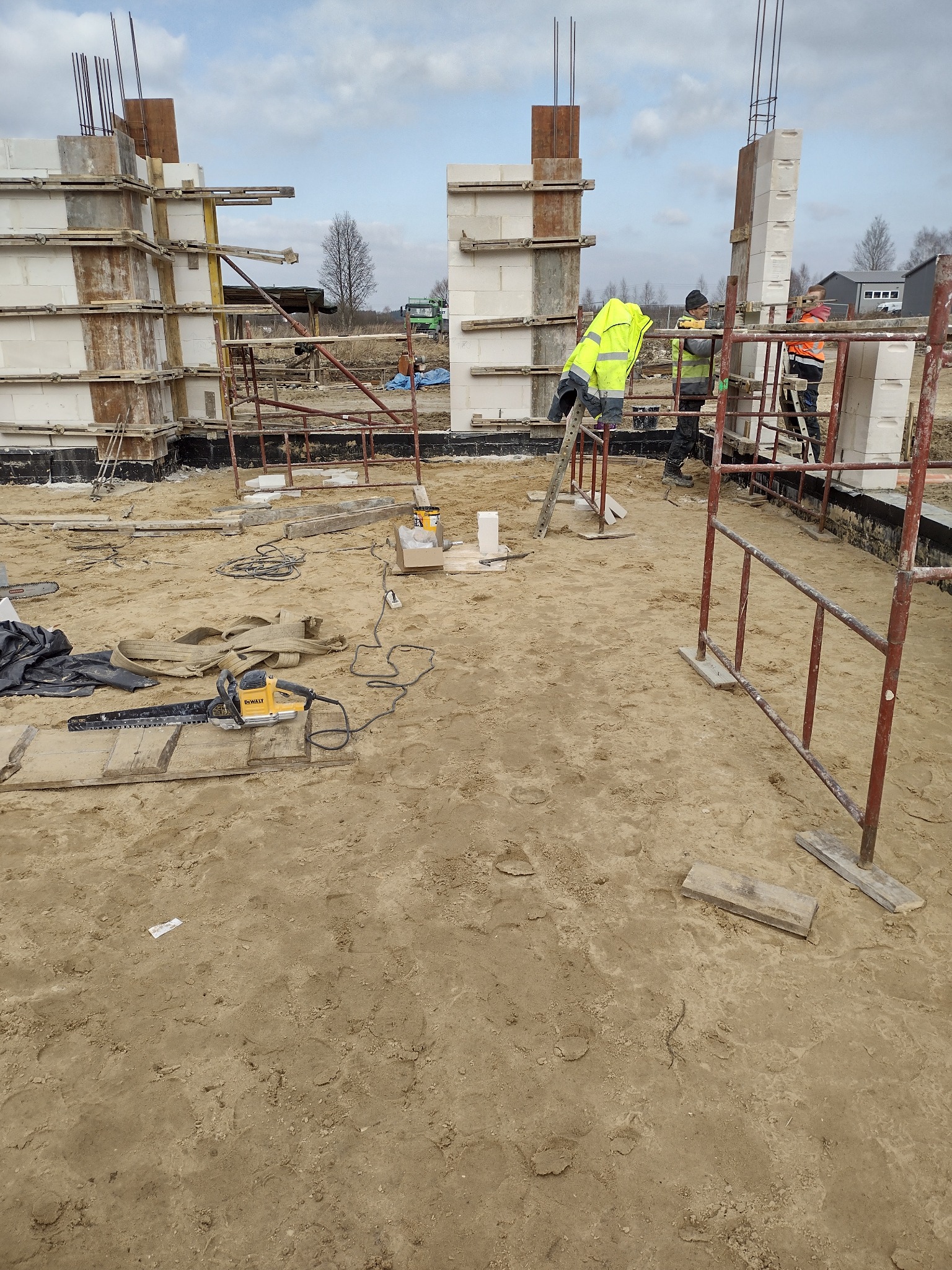 Budowa domu: robotnicy w odblaskowych kamizelkach montują ściany z bloczków na fundamencie, widoczne szalunki, zbrojenia, piła łańcuchowa na ziemi.