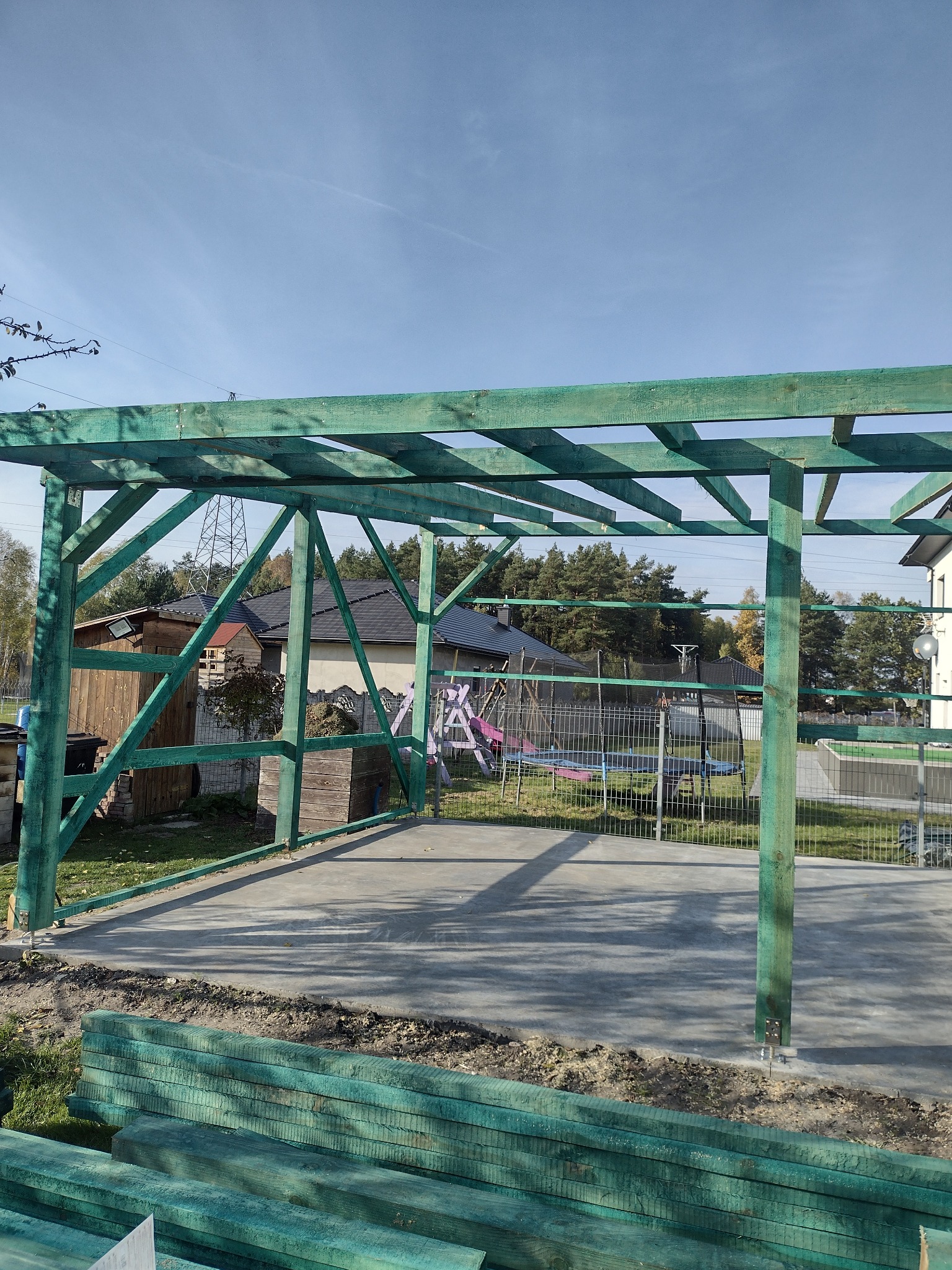 Konstrukcja drewniana, pomalowana na zielono, w trakcie budowy z widocznym betonowym podłożem i stosem belek w tle, w słoneczny dzień.