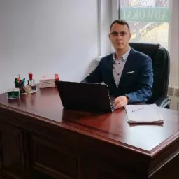 Młody mężczyzna w garniturze siedzi za biurkiem z laptopem, dokumentami i przyborami biurowymi, widoczne okno z napisem na szybie.