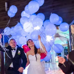 Uśmiechnięta para młoda trzymająca balony w kolorze błękitu i czerwieni, wychodząca z budynku, w tle kobieta w garniturze trzymająca zimne ognie.