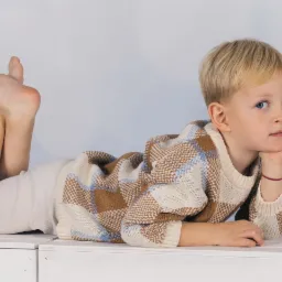 Chłopiec w beżowym swetrze w kratę i spodniach leży na białym podeście, opierając głowę na dłoni, z uniesionymi stopami, na jasnym tle.