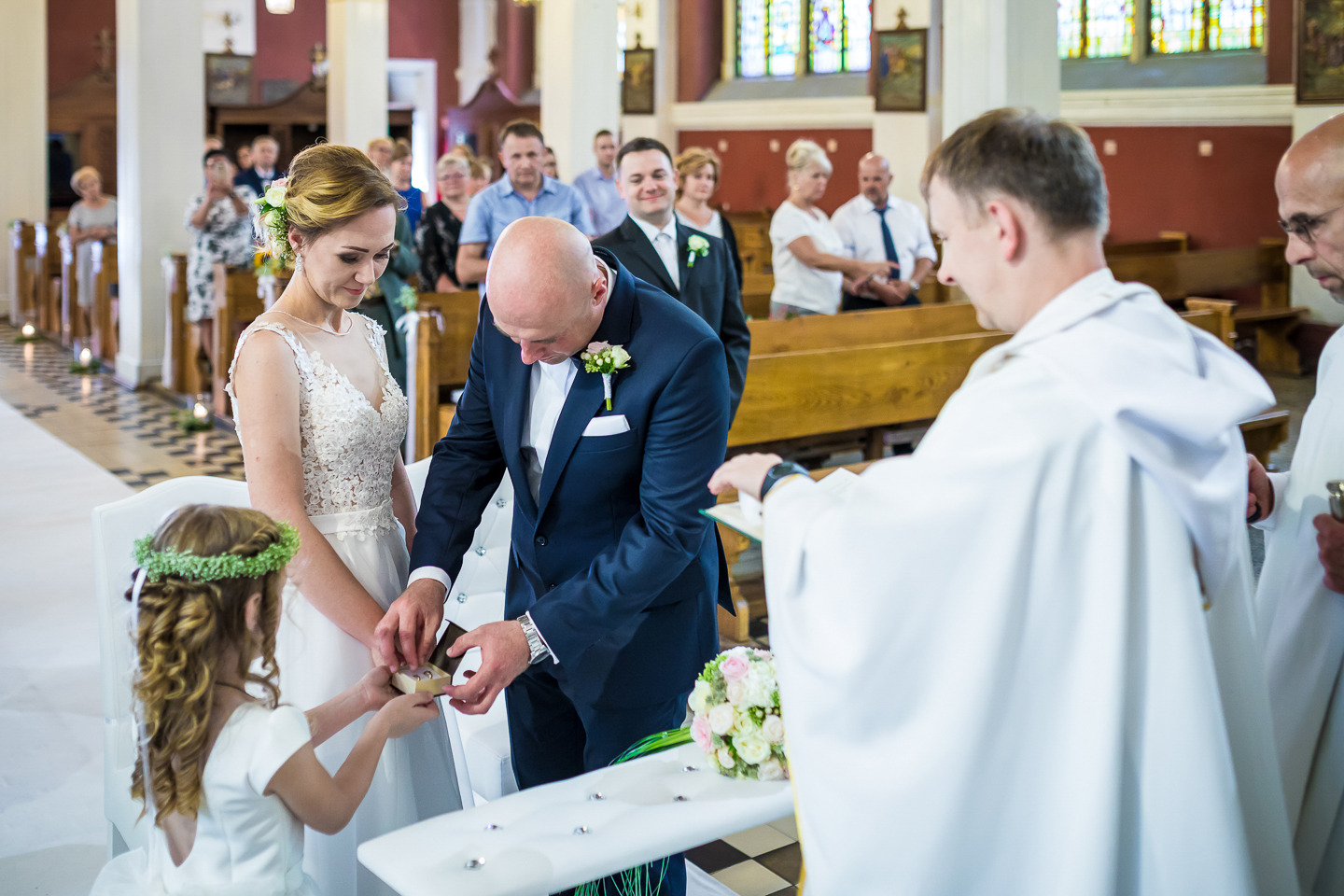 Para młoda wymienia się obrączkami podczas ceremonii ślubnej w kościele, z dziewczynką w wianku podającą pudełko z obrączkami, a księdzem błogosławiącym. W tle goście siedzą w ławkach.