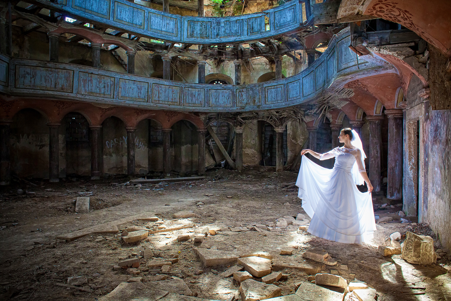 Panna młoda w białej sukni pozuje w ruinach kościoła z drewnianą galerią i kolumnami, światło słoneczne pada na gruz na podłodze.