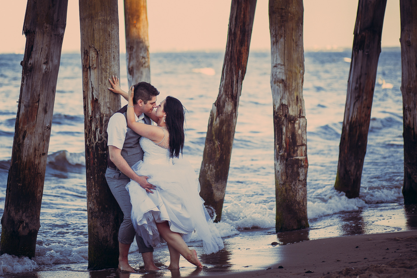 Para młoda w objęciach pod drewnianym molo na plaży, panna młoda w białej sukni, pan młody w szarej kamizelce i spodniach w kratę, fale rozbijają się o filary.