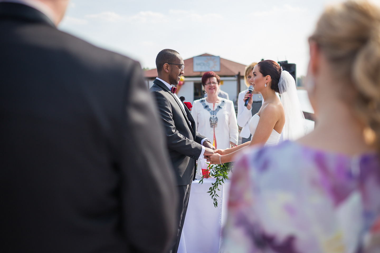 Para młoda trzymająca się za ręce podczas ceremonii ślubnej na zewnątrz, z gośćmi i budynkiem w tle, widok zza ramienia gościa w kwiatowej sukience.