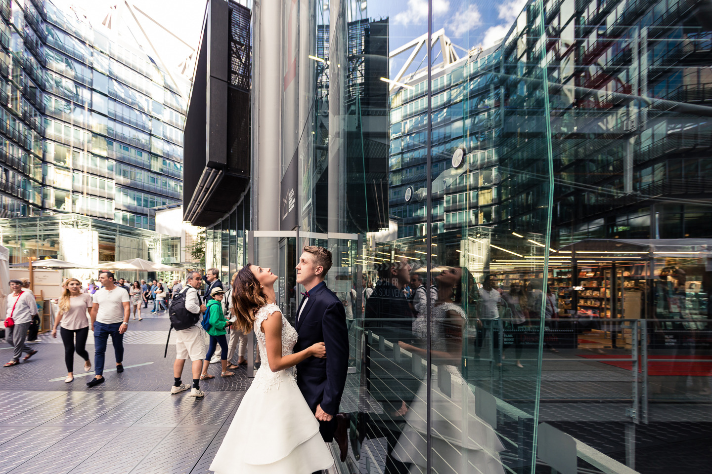 Para młoda w nowoczesnej scenerii miejskiej: panna młoda w białej sukni z koronkową górą i pan młody w granatowym garniturze, oboje patrzą w górę, odbijając się w szklanej fasadzie budynku, w tle...
