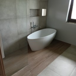 Mateusz Pindel Wykończenia Wnętrz - Nowoczesna łazienka z wolnostojącą białą wanną, szarymi płytkami wielkoformatowymi na ścianie z dekoracyjną wnęką i drewnopodobną podłogą, widok na szklaną ścianę prysznicową i okno.