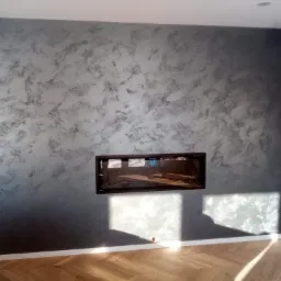 Ściana z dekoracyjną szarą gładzią w salonie, z wbudowanym kominkiem i parkietem ułożonym w jodełkę, oświetlona promieniami słońca.