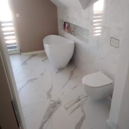 Mateusz Pindel Wykończenia Wnętrz - Nowoczesna łazienka na poddaszu z białą, wolnostojącą wanną, toaletą i marmurowymi płytkami. W ścianie wnęka z kosmetykami. Jasne, naturalne światło.