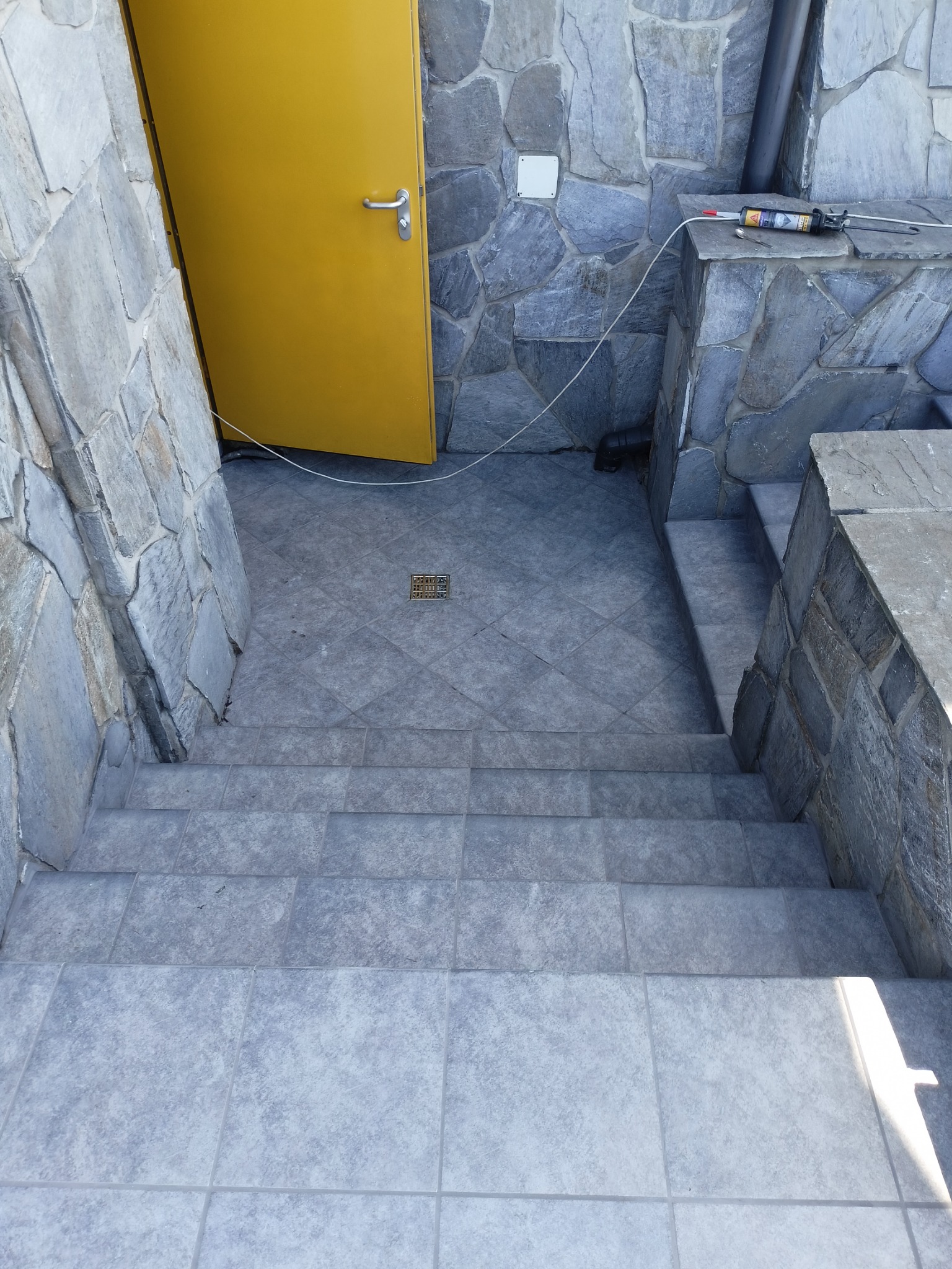 Schody zewnętrzne wyłożone szarymi płytkami, prowadzące do żółtych drzwi, z kamienną ścianą po bokach i tubą silikonu na murku.