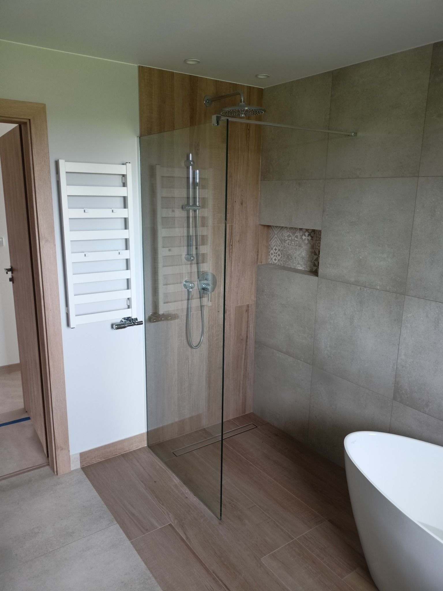 Nowoczesna łazienka z kabiną prysznicową typu walk-in, grzejnikiem drabinkowym na ścianie, i wolnostojącą wanną, wykończona płytkami w odcieniach beżu i szarości, z akcentem dekoracyjnej niszy.