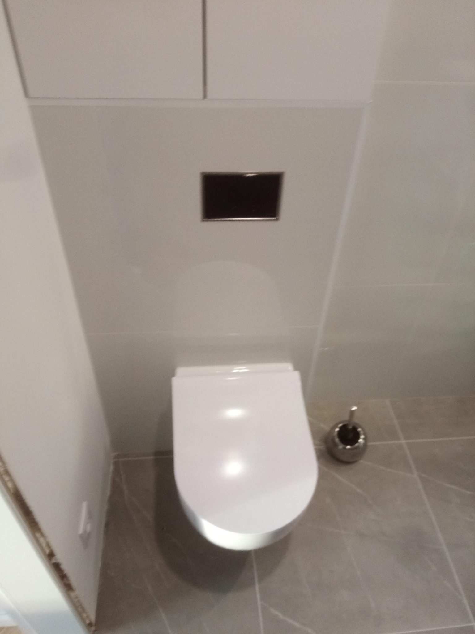 Nowoczesna toaleta podwieszana w jasnej, wykafelkowanej łazience z szarą podłogą i zabudową nad nią. Minimalistyczny styl i czyste linie.