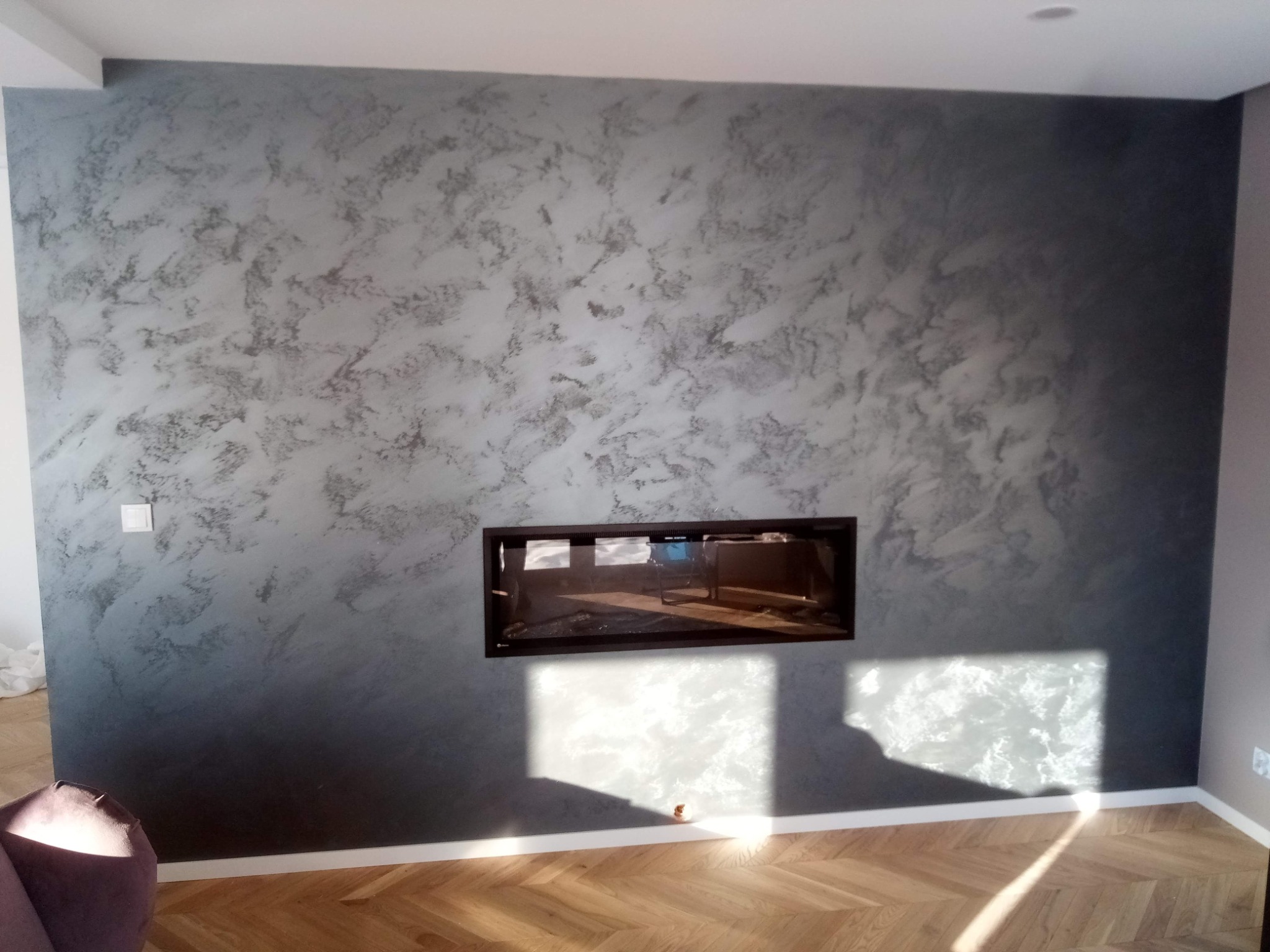 Ściana z dekoracyjną szarą gładzią w salonie, z wbudowanym kominkiem i parkietem ułożonym w jodełkę, oświetlona promieniami słońca.