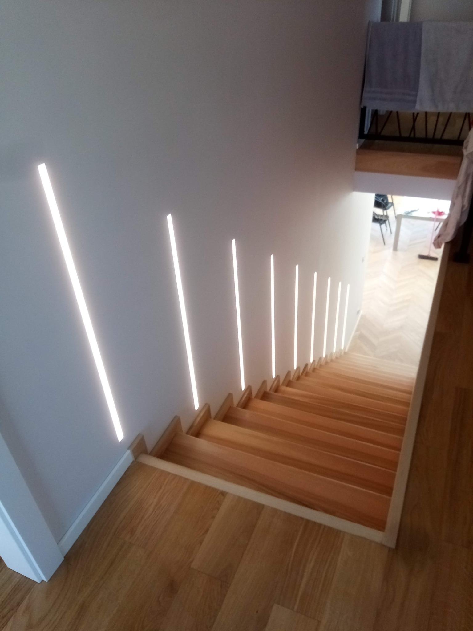 Nowoczesne schody drewniane z liniowym oświetleniem LED wzdłuż ściany. Parkiet w jodełkę widoczny na podłodze. Minimalistyczny design.