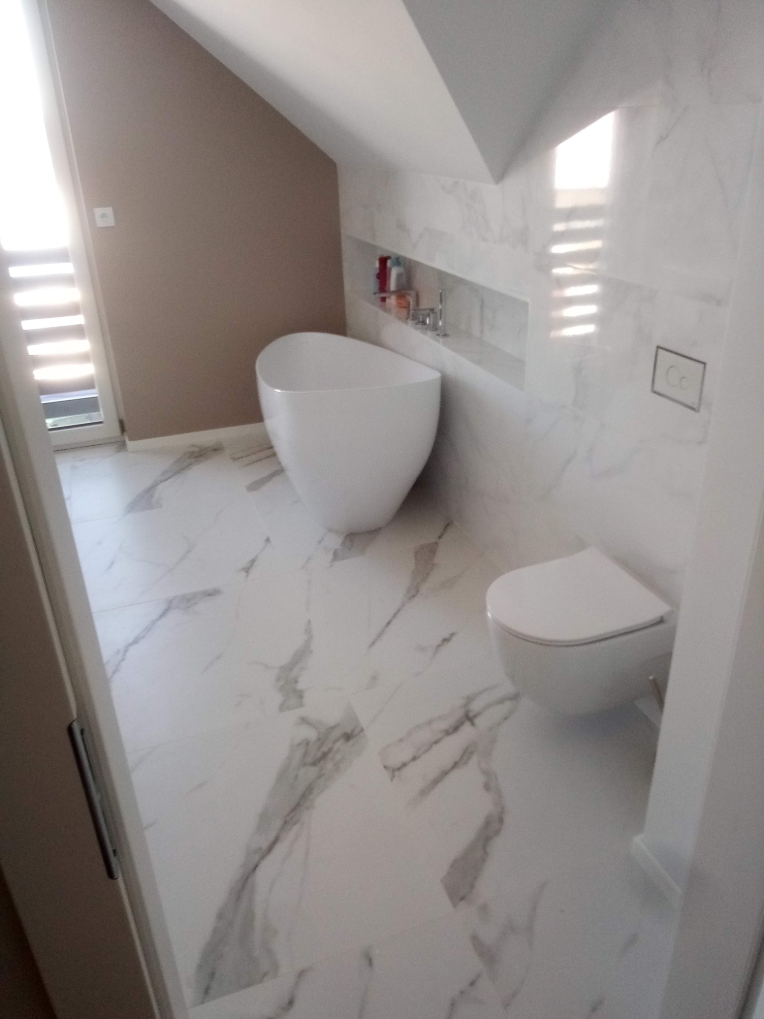 Nowoczesna łazienka na poddaszu z białą, wolnostojącą wanną, toaletą i marmurowymi płytkami. W ścianie wnęka z kosmetykami. Jasne, naturalne światło.