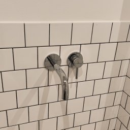 ForosKY Sp.z o.o. - Bateria umywalkowa chromowana, zamontowana na ścianie z białych płytek z czarną fugą. Widoczne pudełka po płytkach. Minimalistyczny design.