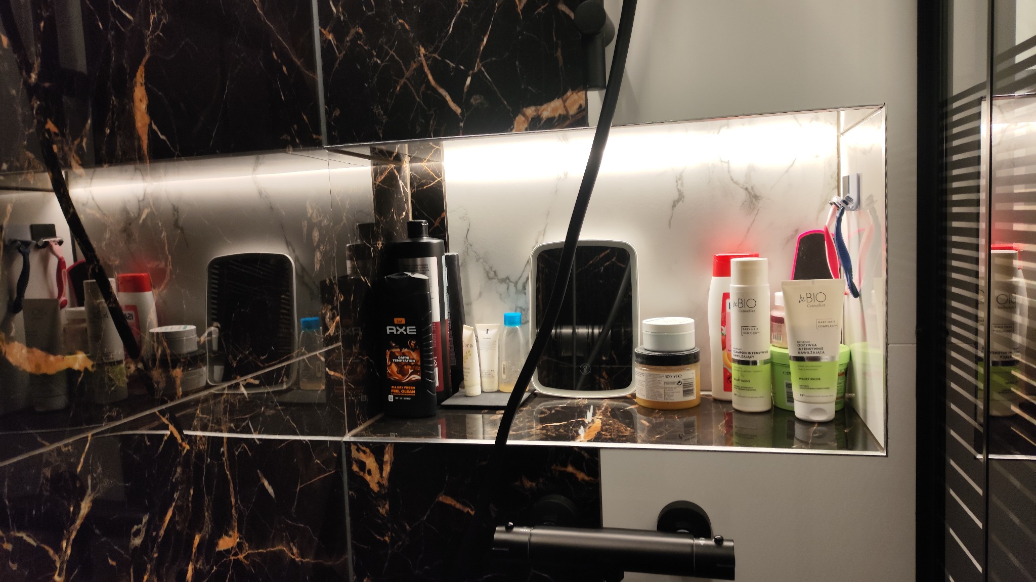 Półka w łazience z czarnymi marmurowymi płytkami, oświetlona listwą LED, na której stoją kosmetyki i akcesoria do higieny osobistej, w tym produkty marek Axe i BeBio.